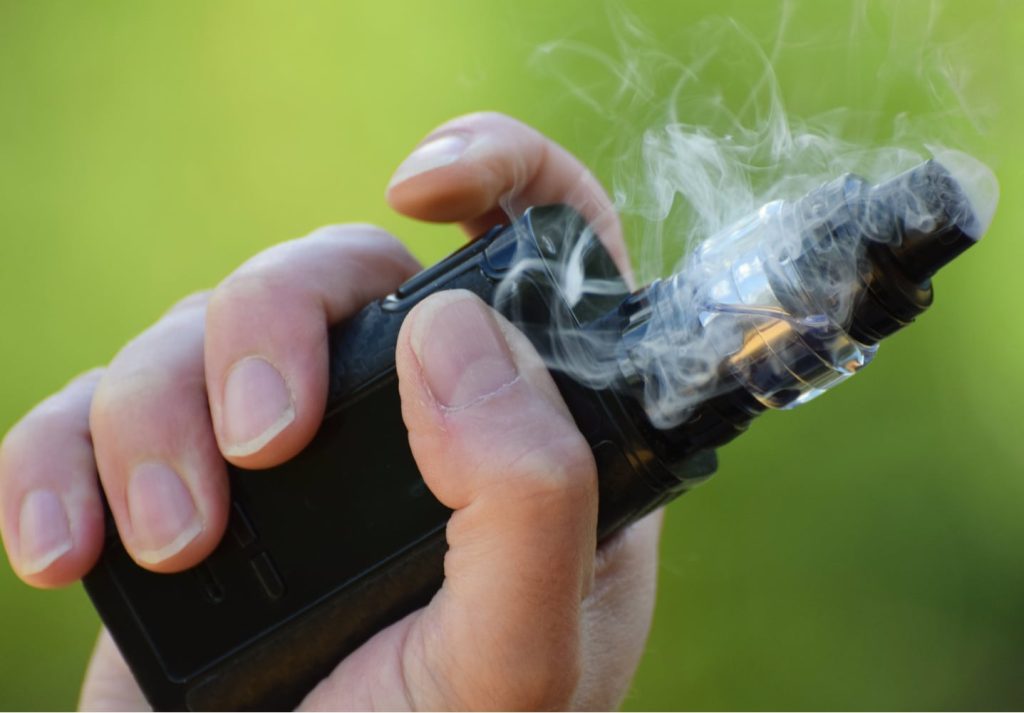 Mão segurando um vape com fumaça saindo do bico do equipamento