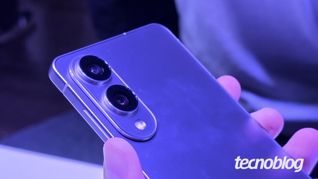 Imagem colorida mostra um smartphone Samsung Galaxy S25 Edge sendo segurado em uma m&atilde;o, com a traseira para cima. A foto est&aacute; em close-up nas duas c&acirc;meras traseiras. Na parte inferior direita, o logotipo do "Tecnoblog" &eacute; vis&iacute;vel.