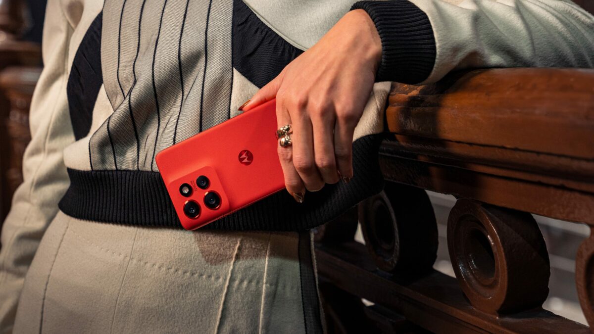Smartphone Motorola vermelho em destaque, exibindo tr&ecirc;s c&acirc;meras traseiras e o logotipo da marca no centro. O aparelho &eacute; segurado por uma pessoa encostada em um corrim&atilde;o de madeira, com roupa clara e detalhes escuros.