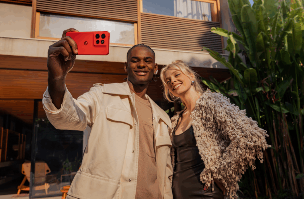 Duas pessoas posam para uma selfie ao ar livre enquanto uma delas segura um smartphone Motorola vermelho, voltado para a c&acirc;mera. O aparelho exibe tr&ecirc;s lentes traseiras e o logotipo da marca. A dupla sorri em frente a uma casa moderna, com plantas tropicais ao fundo e luz natural destacando o design vibrante do celular.