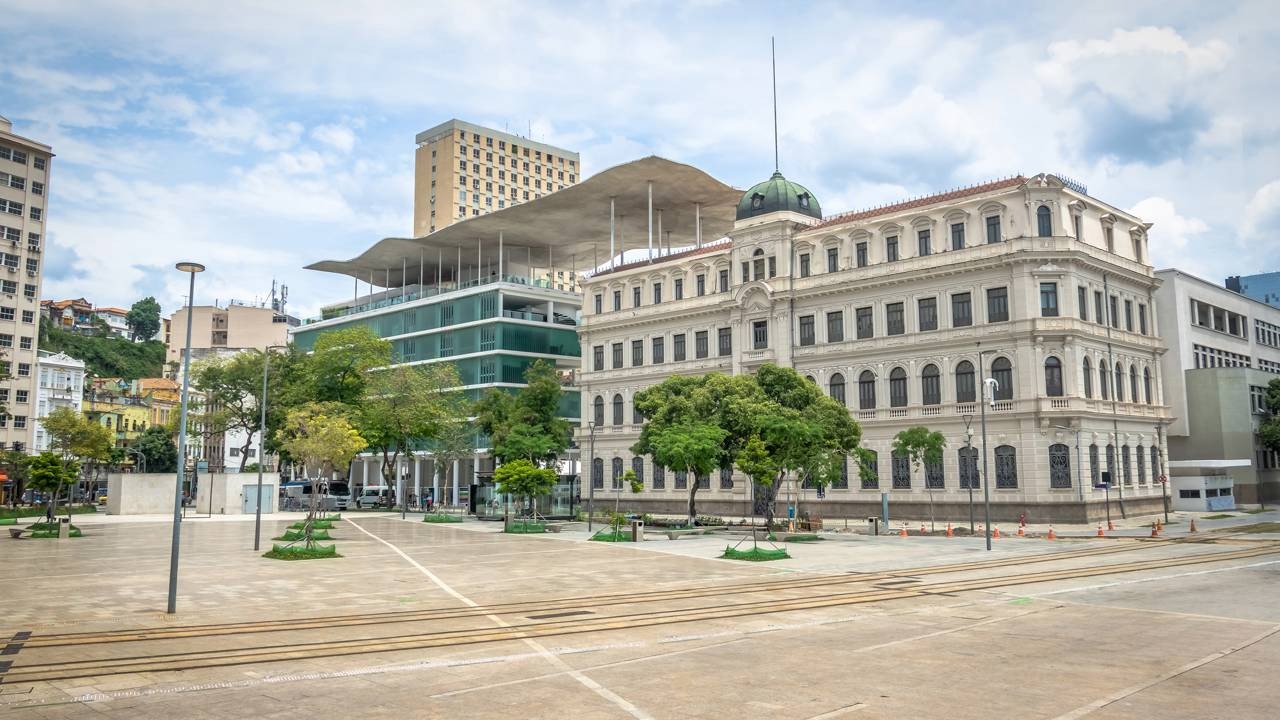 Praça urbana no Rio de Janeiro com prédios históricos e modernos, árvores e trilhos no chão.