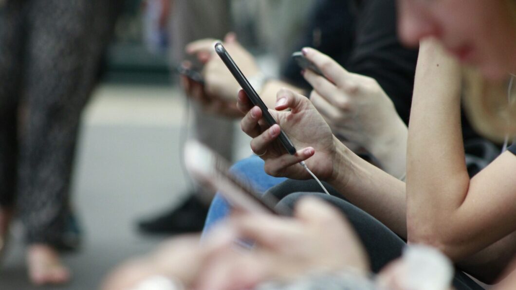 Foto de pessoas sentadas usando smartphones. O foco da imagem são os smartphones, e as pessoas não aparecem.