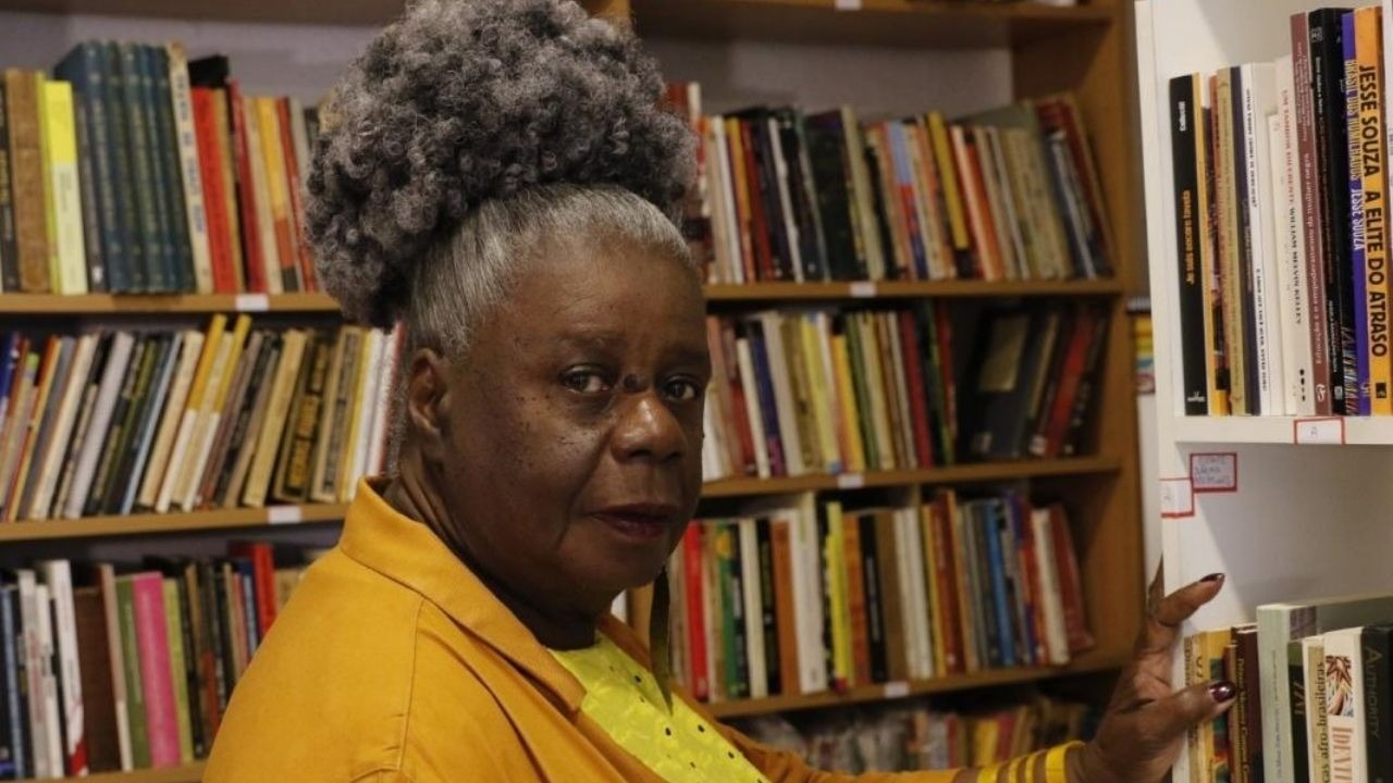 Conceição Evaristo em livraria, cabelo crespo, blusa amarela, tocando livro na prateleira