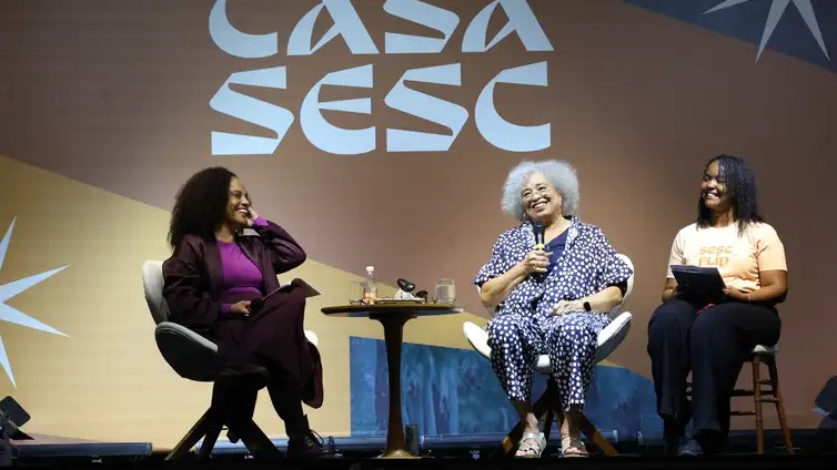 Três mulheres sorrindo em palco com "CASA SESC": uma segura microfone, outra tablet