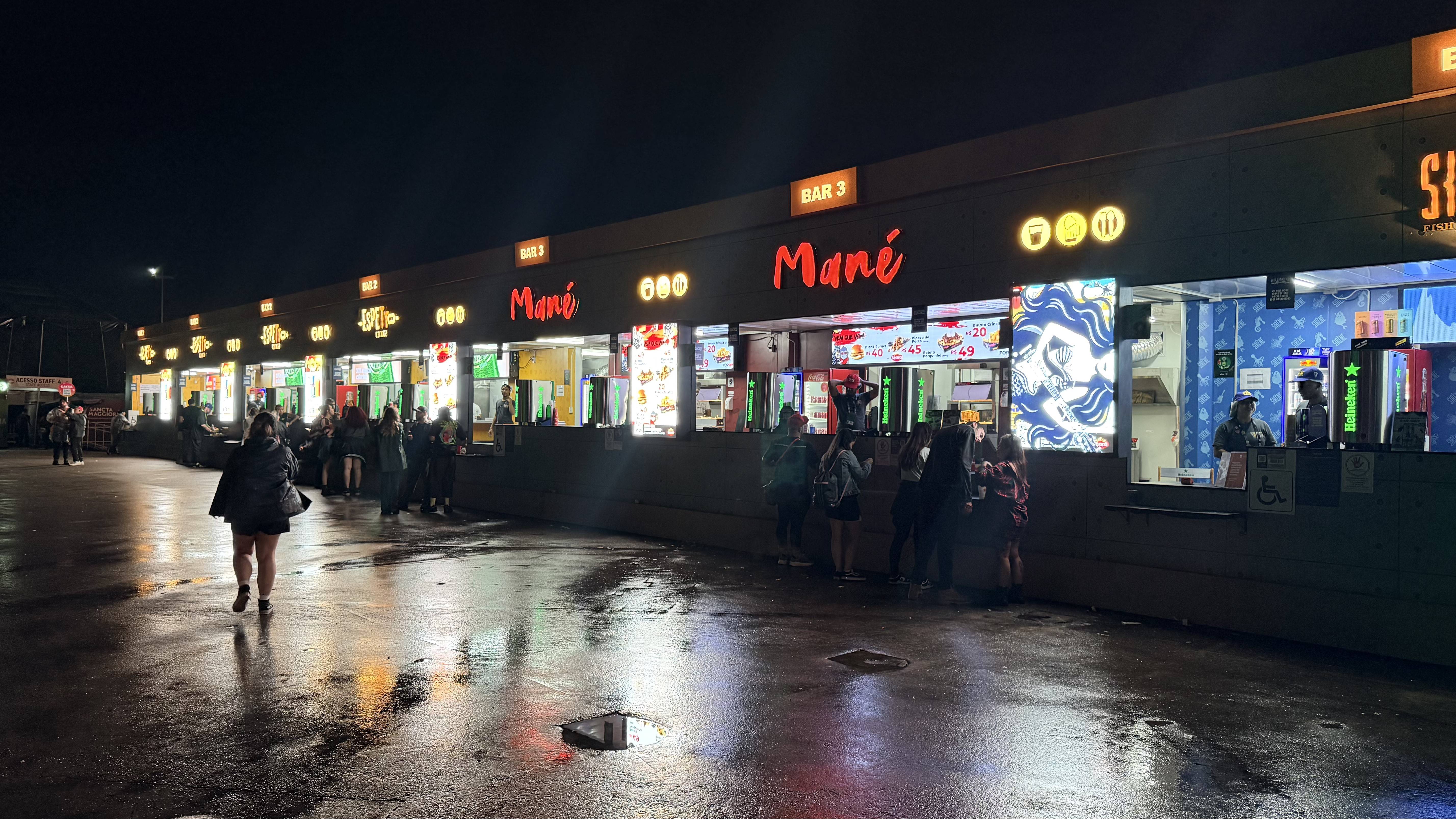 Pessoas em frente a uma fileira de barracas de comida e bebida iluminadas à noite, com o chão molhado refletindo as luzes. As barracas têm letreiros Mané em vermelho e Bar 3 em laranja.